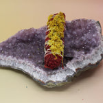 The Organic Dragon's Blood White Sage with Wildflowers - Protection and Love 4 in. Smudge Stick on an amethyst piece all on a pink and yellow background.