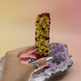 Someone holding the Organic Dragon's Blood White Sage with Wildflowers - Protection and Love 4 in. Smudge Stick in front of an amethyst piece all on a pink and yellow background.