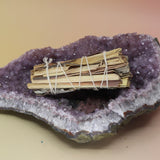 The Organic White Sage & Lemongrass - Clarity and Focus 4 in. Sage Smudge Stick on an amethyst piece all on a pink and yellow background.