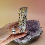 Someone holding Organic White Sage & Lemongrass - Clarity and Focus 4 in. Sage Smudge Stick in front of an amethyst piece on a pink and yellow background.