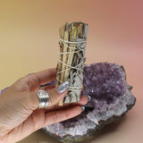 Someone holding Organic White Sage & Lemongrass - Clarity and Focus 4 in. Sage Smudge Stick in front of an amethyst piece on a pink and yellow background.
