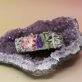 The 'Fairy Dreams' White Sage with Mixed Flowers - 4 in. Smudge Stick on an amethyst piece all on a pink and yellow background.