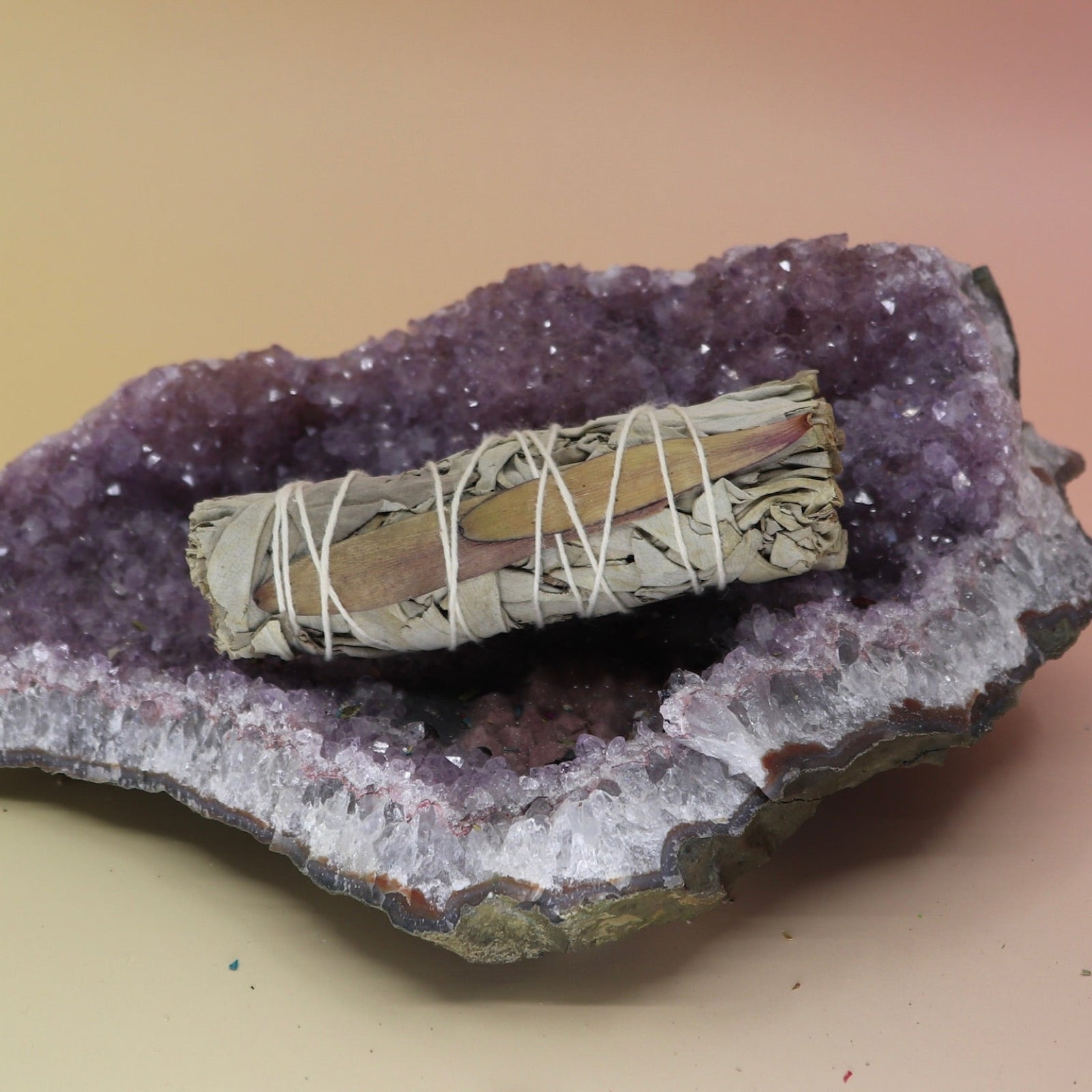 The 'Dream' Organic Floral White Sage with Flowers - 4 in. Smudge Stick on an amethyst piece all on a pink and yellow background.