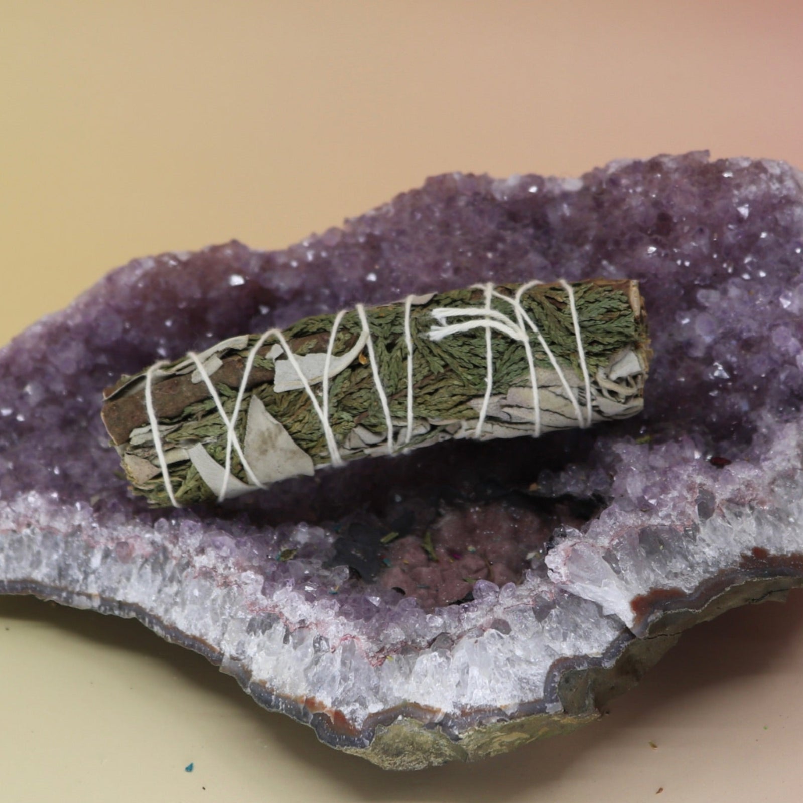 The Organic White Sage & Cedar - 4 in. Purifying and Balancing Smudge Stick on an amethyst piece all on a pink and yellow background.