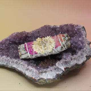 The 'Pristine Pink' Organic White Sage with Statice Flower and Rose Petals - 4 in. Smudge Stick on an amethyst piece on a pink and yellow background.