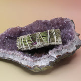 The 'Good Life' Organic Floral White Sage - 4 in. Sage Smudge Stick on an amethyst piece all on a pink and yellow background.