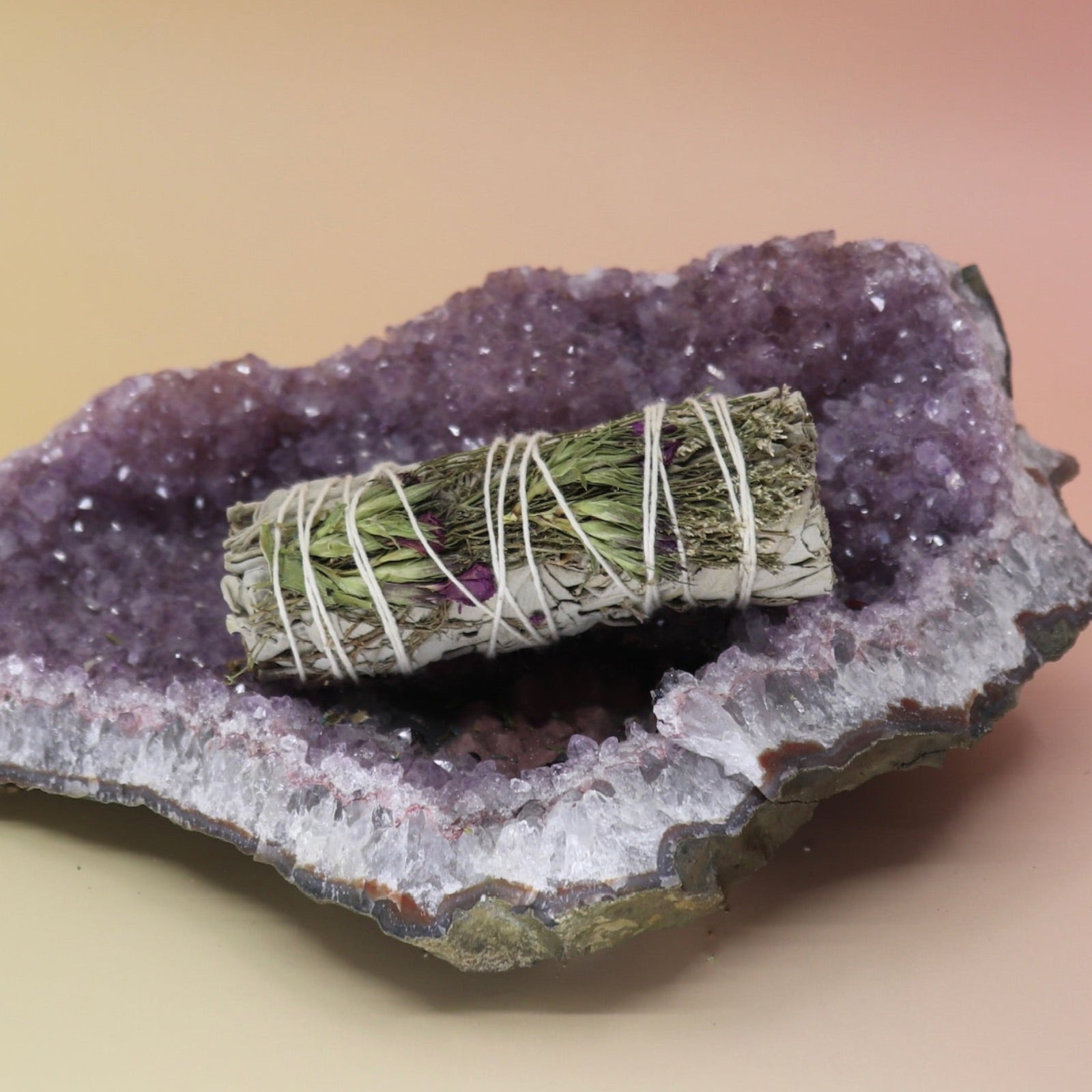 The 'Good Life' Organic Floral White Sage - 4 in. Sage Smudge Stick on an amethyst piece all on a pink and yellow background.