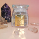 The Royal Frankincense in front of two crystal pieces with crystal points surrounding the jar all on a pink and yellow background.