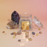 The Royal Frankincense jar with its lid off, in front of two crystal pieces with frankincense surrounding the jar all on a pink and yellow background.