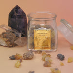 The Royal Frankincense jar with its lid off, in front of two crystal pieces with frankincense surrounding the jar all on a pink and yellow background.