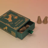 The Mayan Copal + Desert Sage Incense Cones box lying down with two cones next to it all on a pink and yellow background.