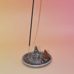 The Silver Buddha Cone and Stick Incense Holder on a pink and yellow background an incense cone and an incense stick on it.