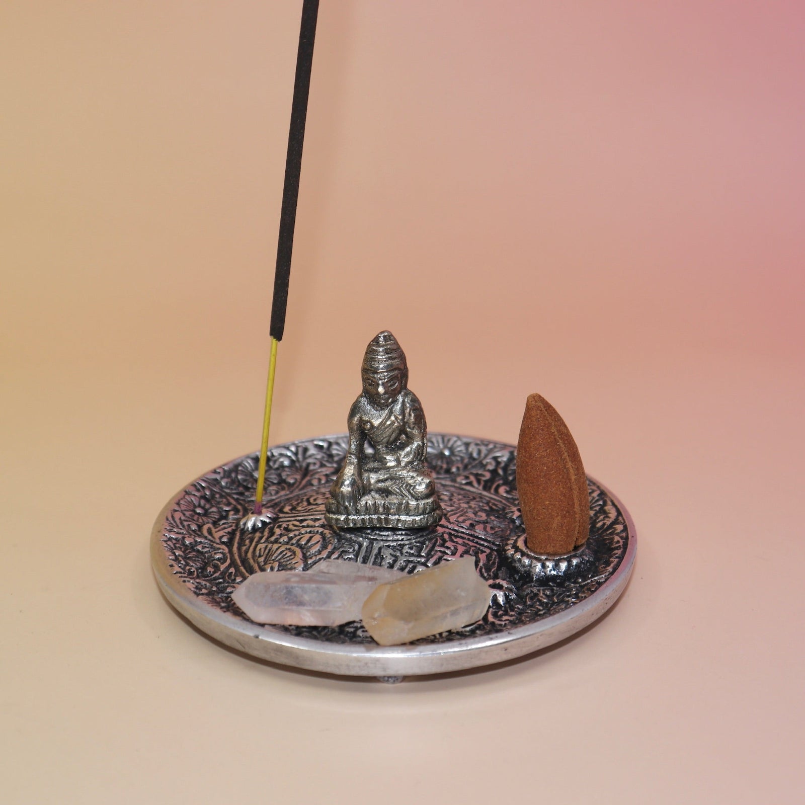 The Silver Buddha Cone and Stick Incense Holder on a pink and yellow background with crystals, an incense cone, and an incense stick on it.