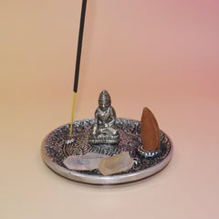 The Silver Buddha Cone and Stick Incense Holder on a pink and yellow background with crystals, an incense cone, and an incense stick on it.