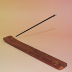 The Om Wooden Incense Burner with an incense stick on a pink and yellow background.