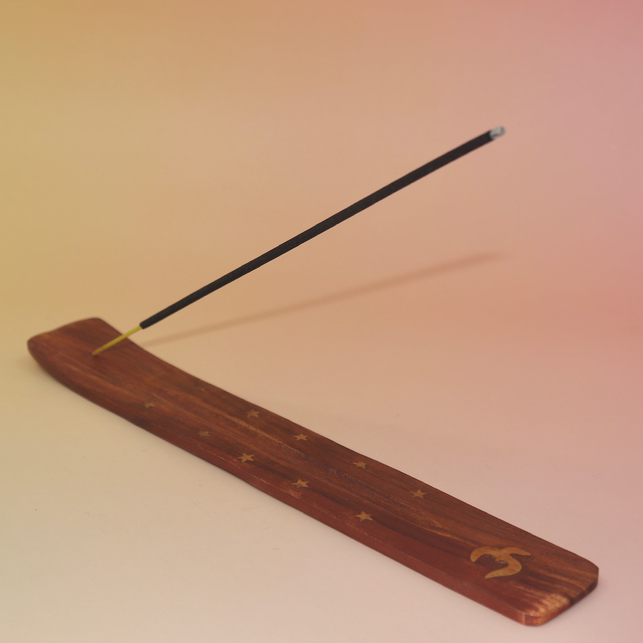 The Om Wooden Incense Burner with an incense stick on a pink and yellow background.