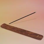 The Moon Wooden Incense Burner with an incense stick on a pink and yellow background.