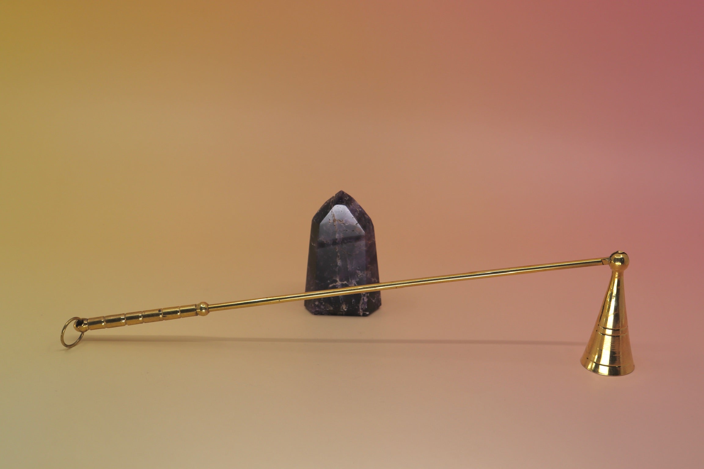 The Brass Candle Snuffer on a pink and yellow background in front on a small is crystal tower. 