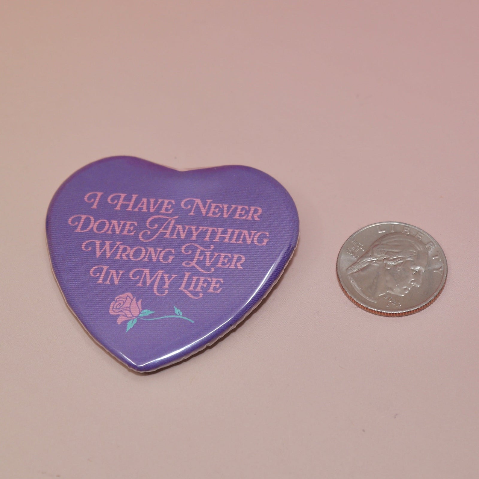 The I Have Never Done Anything Wrong Heart Shaped Pinback Button on a light pink background next to a quarter.