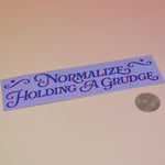The Normalize Holding A Grudge Vinyl Bumper Sticker on a pink and yellow background, next to a quarter.