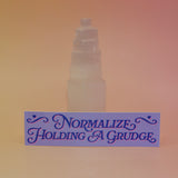 The Normalize Holding A Grudge Vinyl Bumper Sticker against a selenite tower on a pink and yellow background.