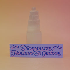 The Normalize Holding A Grudge Vinyl Bumper Sticker against a selenite tower on a pink and yellow background.