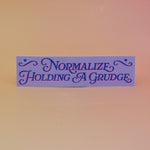 The Normalize Holding A Grudge Vinyl Bumper Sticker on a pink and yellow background.