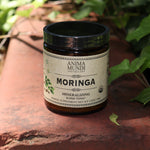 The Moringa Super-Tonic - 4.5oz Powder Energy Booster on red concrete next to greenery.