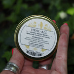 Someone holding the Heal-All Arnica - Nourish, Soothe + Repair Relief Balm in front of ivy.