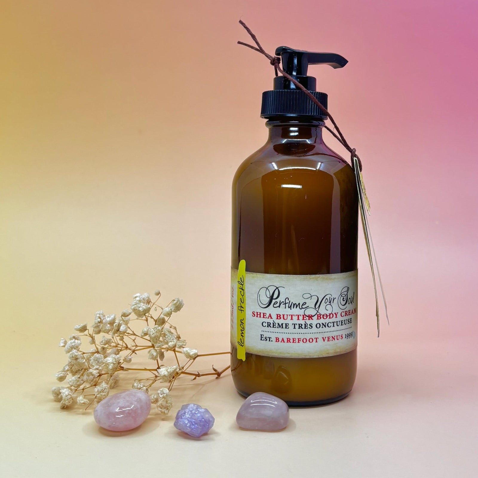The Lemon Freckle Body Cream Moisturizer surrounded by crystals and a dried flower on a light pink background.