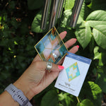 Someone holding the ornament and tag of the Vienna Art Deco Woodstock Wind Chime in front of a bush. 