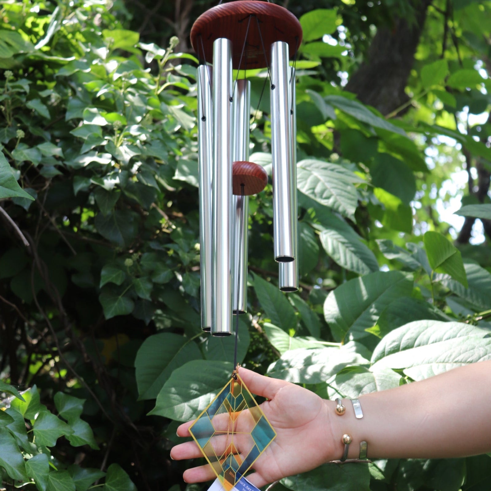 Someone holding the Vienna Art Deco Woodstock Wind Chime in front of a bush. 