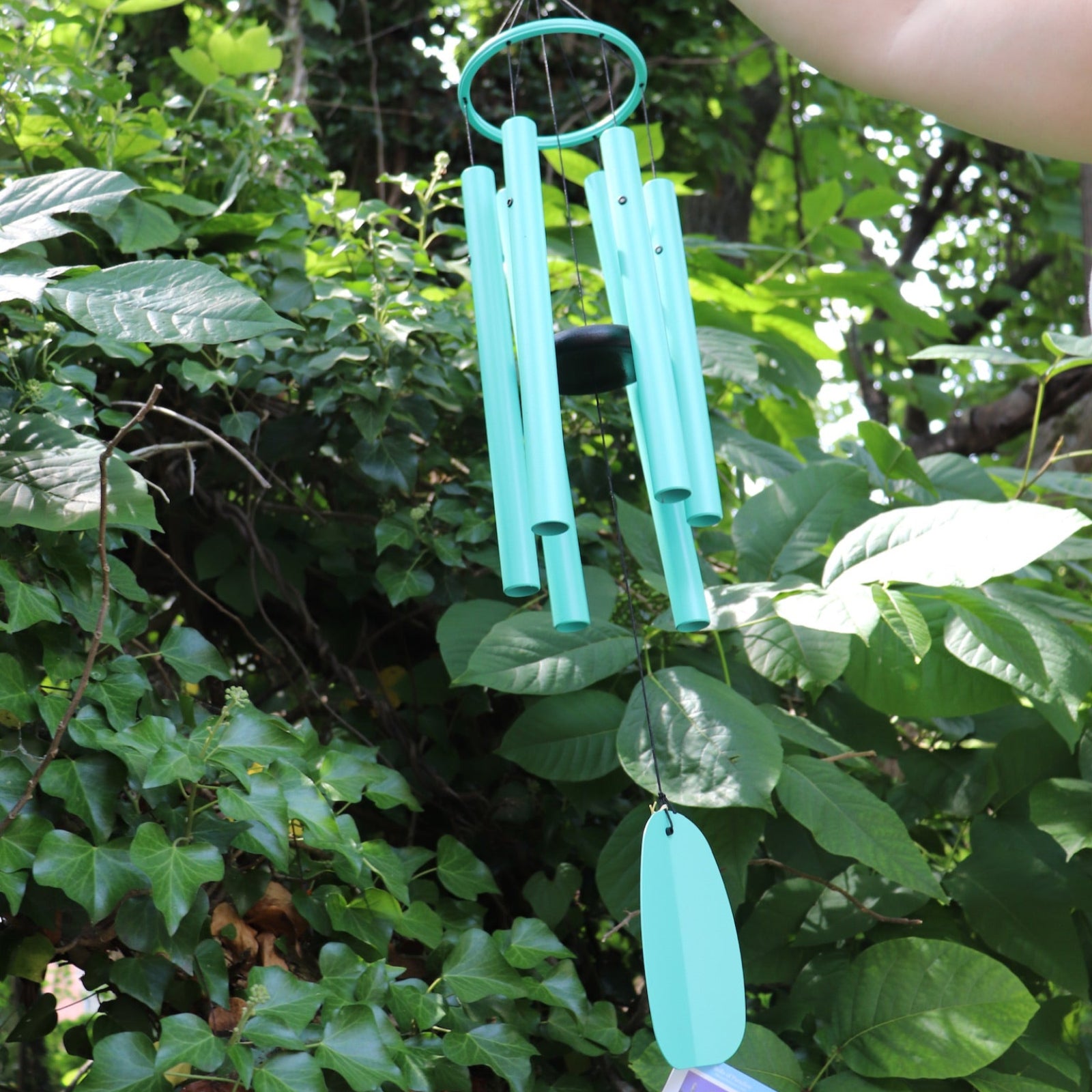 Someone holding the Monochrome Cyan Bells of Paradise Wind Chime in front of a green bush.