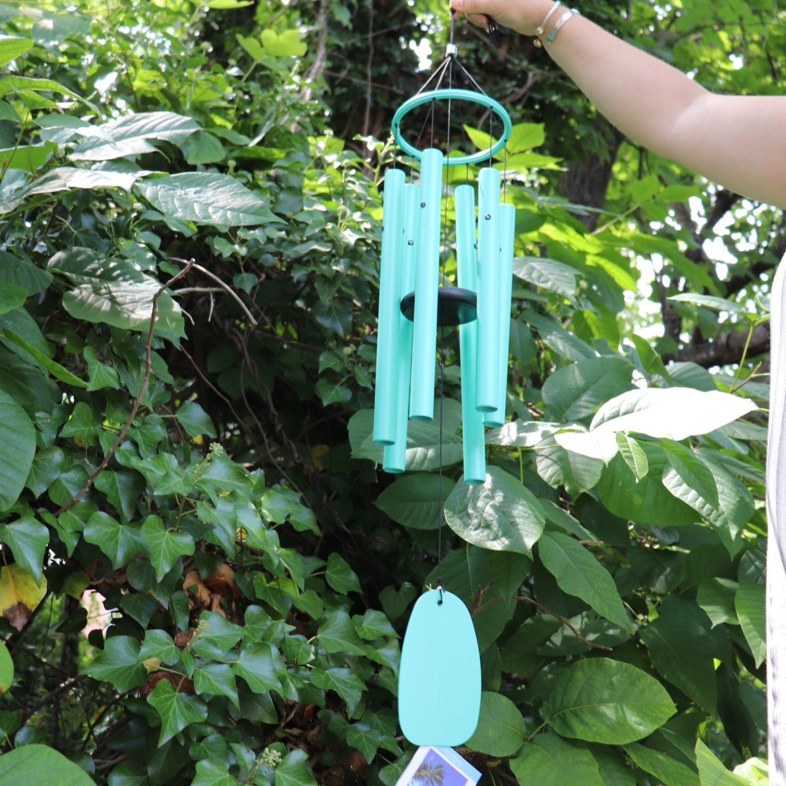Someone holding the Monochrome Cyan Bells of Paradise Wind Chime in front of a green bush.