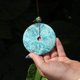 An up-close image of the faux turquoise ornament of the Medium Turquoise Woodstock Wind Chime in front of a green bush.