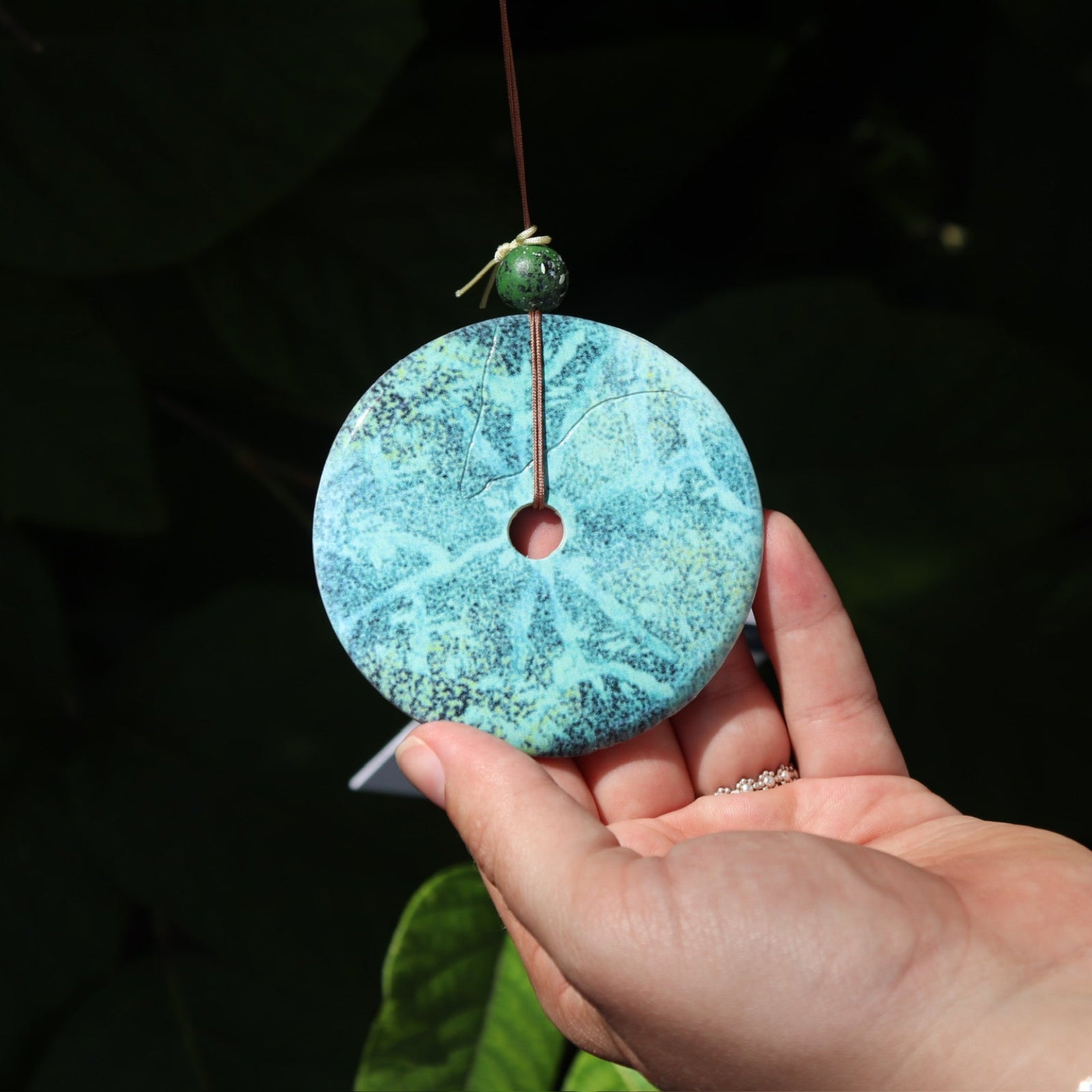 An up-close image of the faux turquoise ornament of the Medium Turquoise Woodstock Wind Chime in front of a green bush.