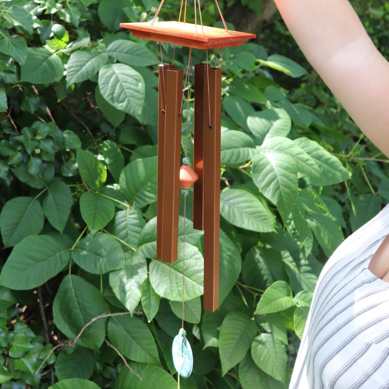 Someone holding the Medium Turquoise Woodstock Wind Chime in front of a green bush.