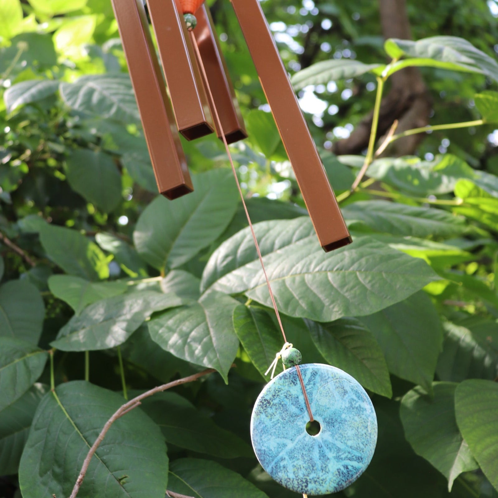 An up-close image of someone holding the Medium Turquoise Woodstock Wind Chime in front of a green bush.