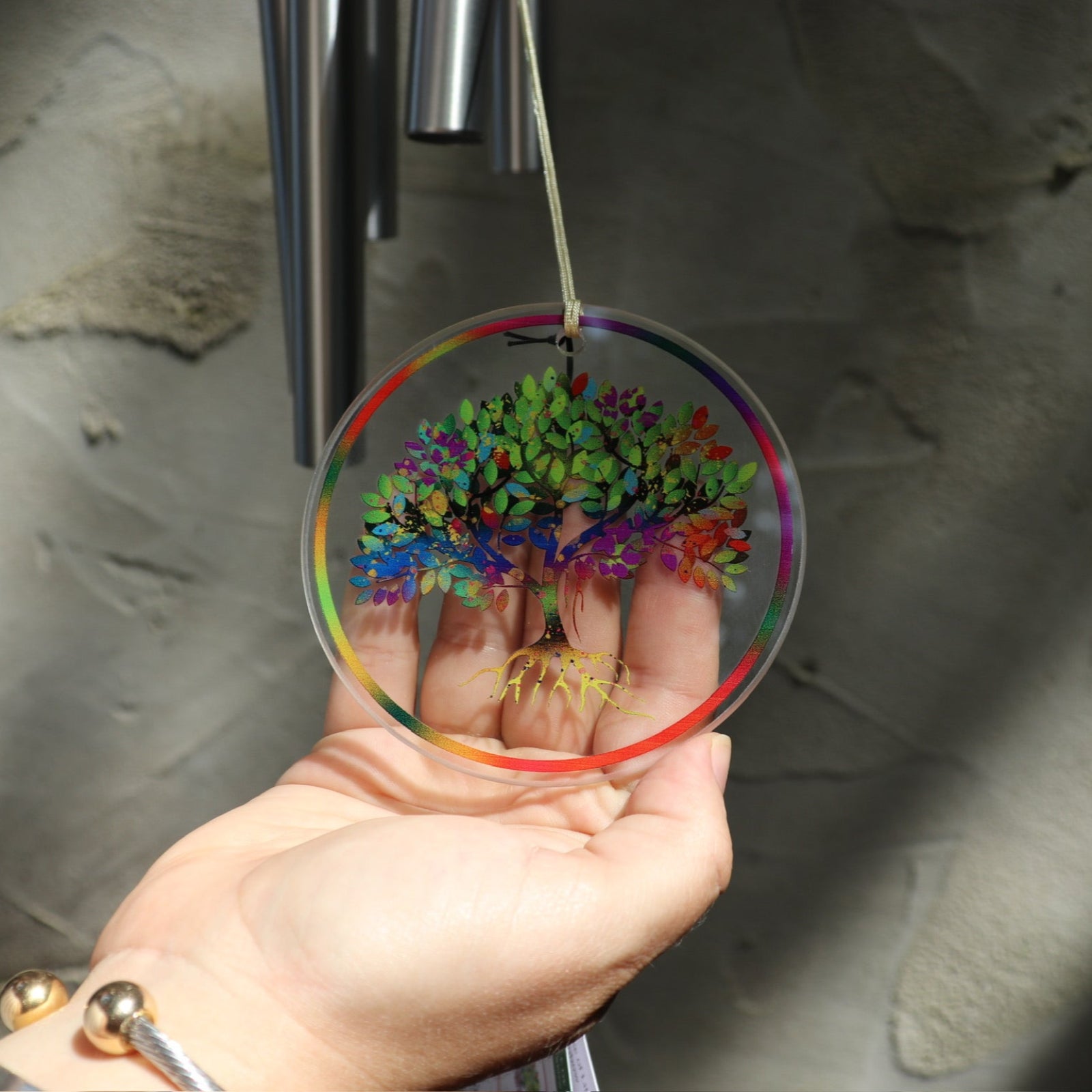 Someone holding the tree of life ornament attached to the Rainbow Tree of Life Woodstock Wind Chime in front of a white wall.