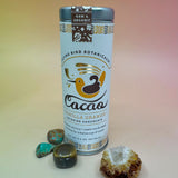 The Vanilla Orange Cacao Small Tin on a pink and yellow background surrounded by small crystals.