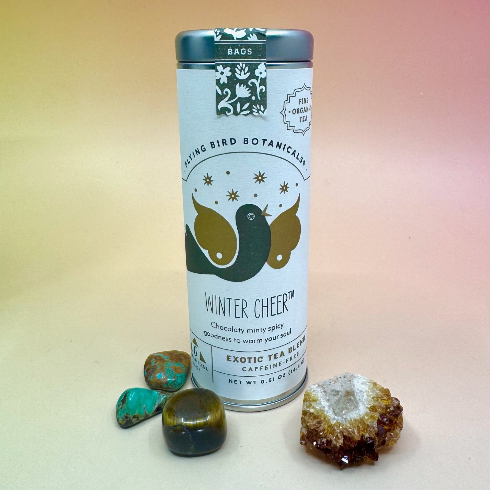 The Winter Cheer 6 Tea Bag Tin on a pink and yellow background surrounded by different small crystals.