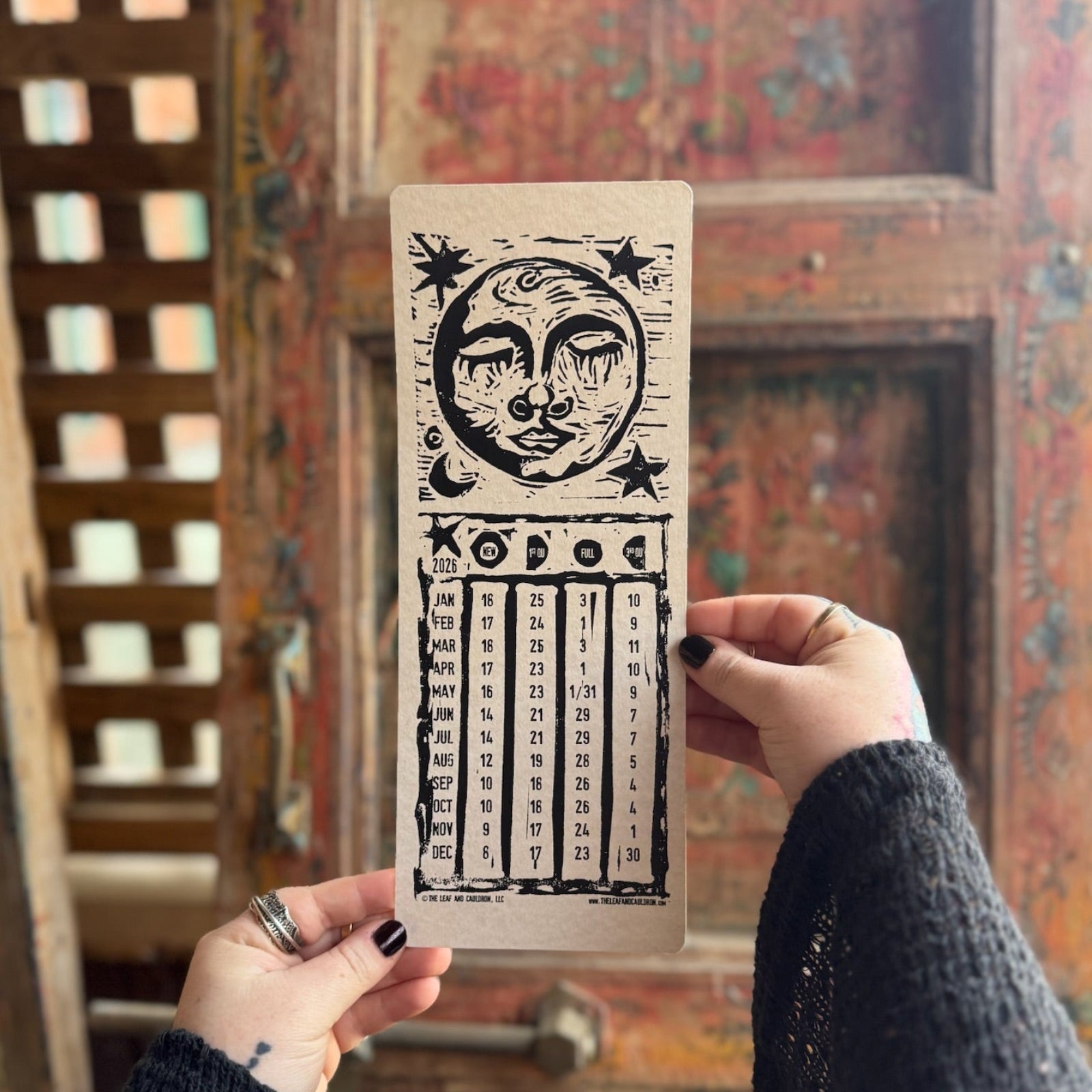 Someone holding the 2026 Moon Phase Calendar in front of a wooden background.