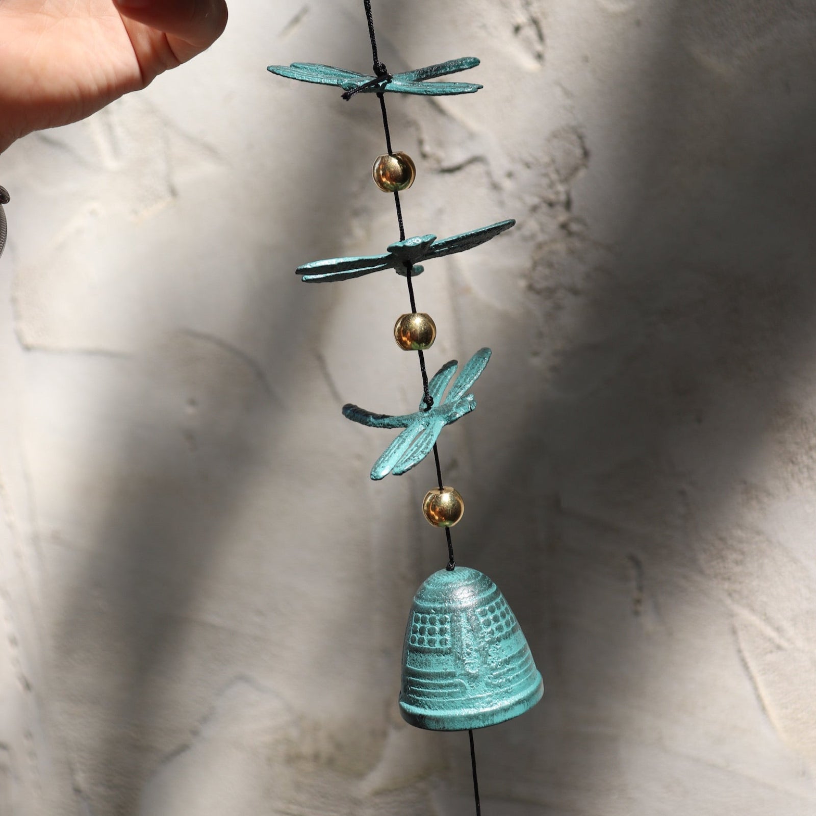 Someone holding the Verdigris Dragonfly Woodstock Habitats Windbell in front of a white concrete wall.