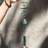 Someone holding the Verdigris Dragonfly Woodstock Habitats Windbell in front of a white concrete wall.