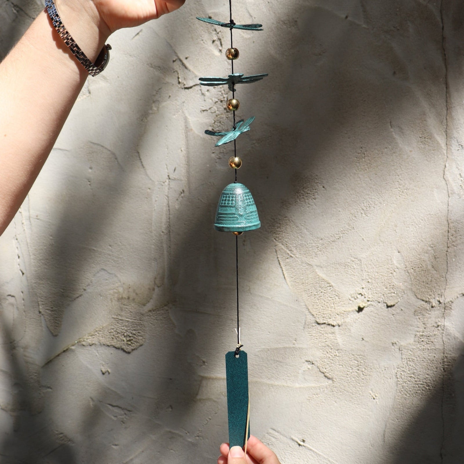 Someone holding the Verdigris Dragonfly Woodstock Habitats Windbell in front of a white concrete wall.