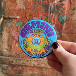 Someone holding the Gemini Astral Weekend Zodiac Magnet 3" Round in front of a wooden background.