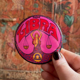 Someone holding the Libra Astral Weekend Zodiac Magnet 3" Round in front of a wooden background.