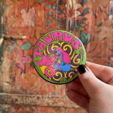 Someone holding the Taurus Astral Weekend Zodiac Magnet 3" Round in front of a wooden background.