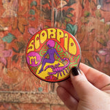 Someone holding the Scorpio Astral Weekend Zodiac Magnet 3" Round in front of a wooden background.