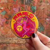 Someone holding the Aries Astral Weekend Zodiac Magnet 3" Round in front of a wooden background.